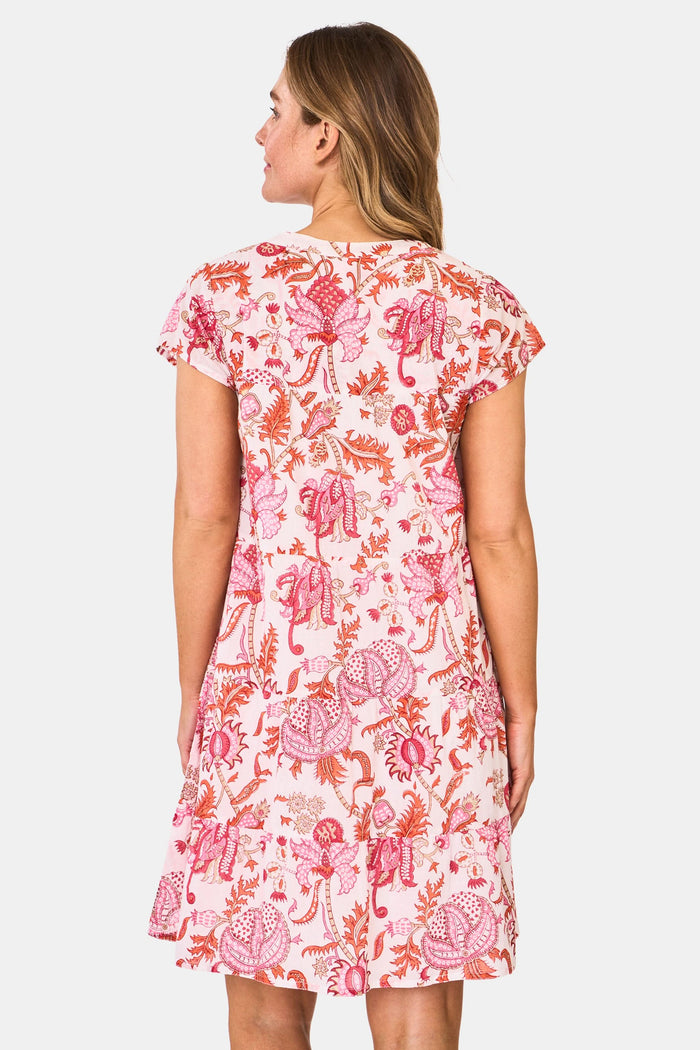 A woman wears the Roller Rabbit Amanda Pamela Dress—a short-sleeved, knee-length, tiered cotton summer dress with a pink and red floral print—standing against a plain light background, smiling with one hand on the hem.