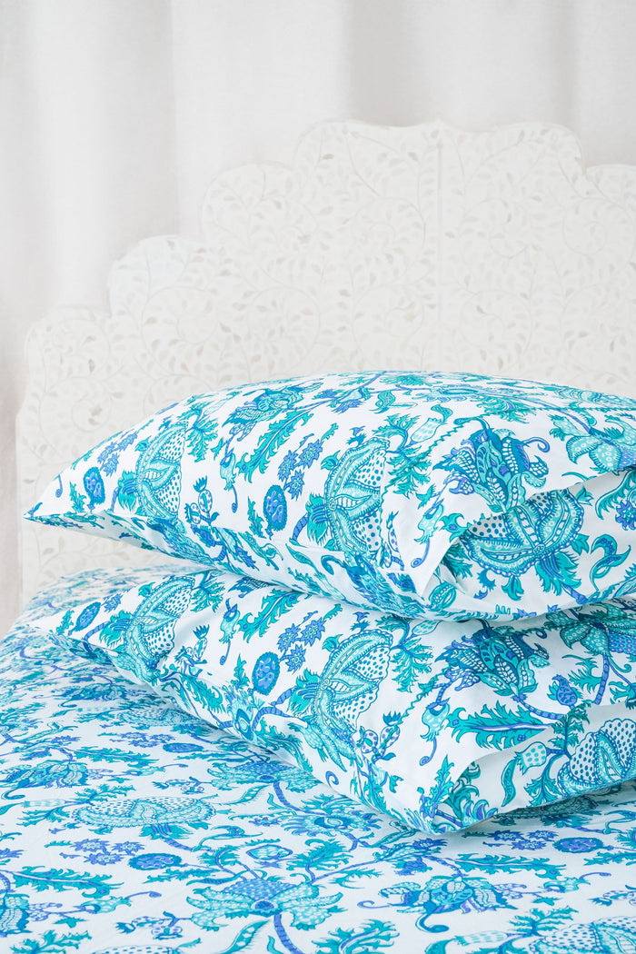 Two pillows with Amanda Pillowcase Set by Roller Rabbit rest on blue and turquoise floral bedding; the 100 cotton pillowcases coordinate with a white, leafy headboard and white curtains in the background.