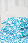 Two pillows with Amanda Pillowcase Set by Roller Rabbit rest on blue and turquoise floral bedding; the 100 cotton pillowcases coordinate with a white, leafy headboard and white curtains in the background.