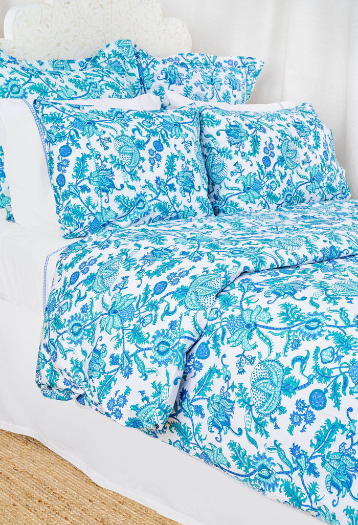 Two pillows with Amanda Pillowcase Set by Roller Rabbit rest on blue and turquoise floral bedding; the 100 cotton pillowcases coordinate with a white, leafy headboard and white curtains in the background.