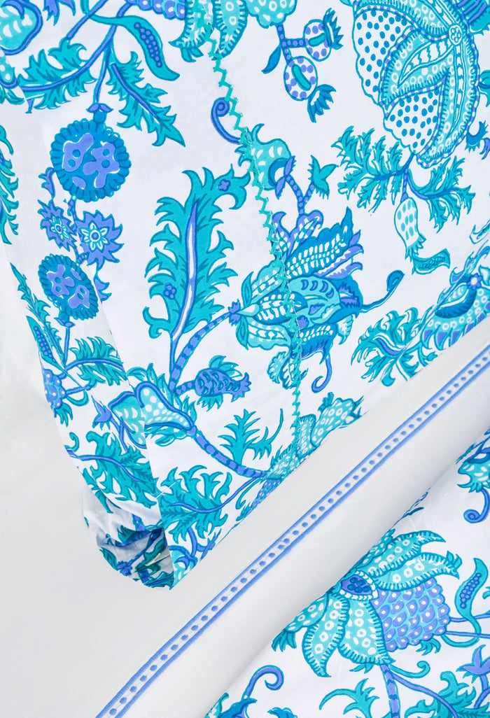 Two pillows with Amanda Pillowcase Set by Roller Rabbit rest on blue and turquoise floral bedding; the 100 cotton pillowcases coordinate with a white, leafy headboard and white curtains in the background.