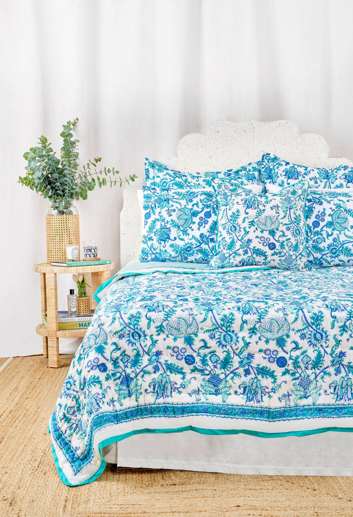 A bed styled with the Amanda Quilt by Roller Rabbit, made from 100 cotton in a blue and white floral design, sits beside a wooden nightstand holding a vase with greenery, books, and a photo frame on a woven rug against a white wall.