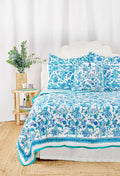 A bed styled with the Amanda Quilt by Roller Rabbit, made from 100 cotton in a blue and white floral design, sits beside a wooden nightstand holding a vase with greenery, books, and a photo frame on a woven rug against a white wall.