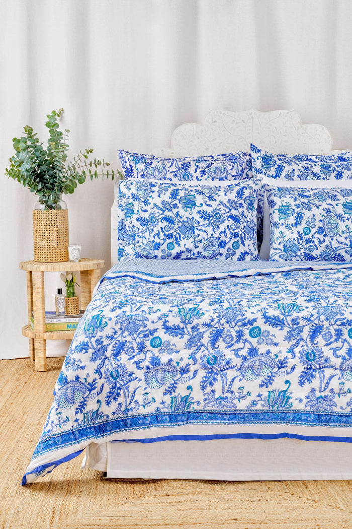 A neatly made bed displays the Roller Rabbit Amanda Quilt, crafted from 100 cotton with a blue and white floral design, set against white curtains. Beside it, a small wooden table holds books, a basket, and a vase on a woven rug.
