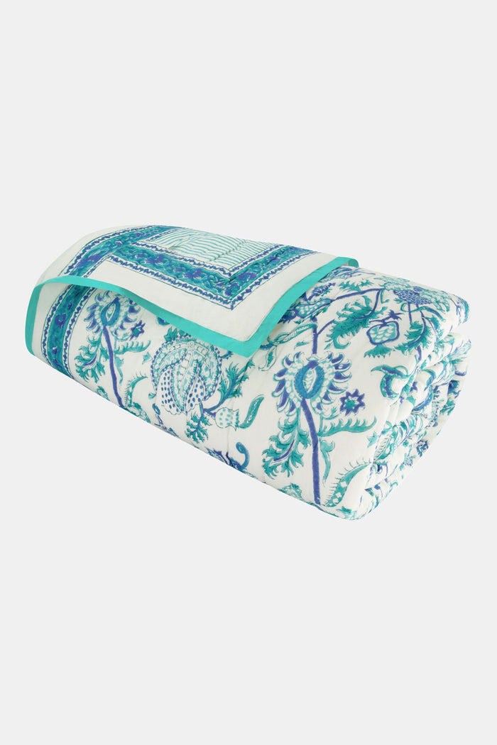 A bed styled with the Amanda Quilt by Roller Rabbit, made from 100 cotton in a blue and white floral design, sits beside a wooden nightstand holding a vase with greenery, books, and a photo frame on a woven rug against a white wall.