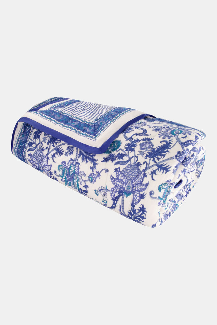 A neatly made bed displays the Roller Rabbit Amanda Quilt, crafted from 100 cotton with a blue and white floral design, set against white curtains. Beside it, a small wooden table holds books, a basket, and a vase on a woven rug.