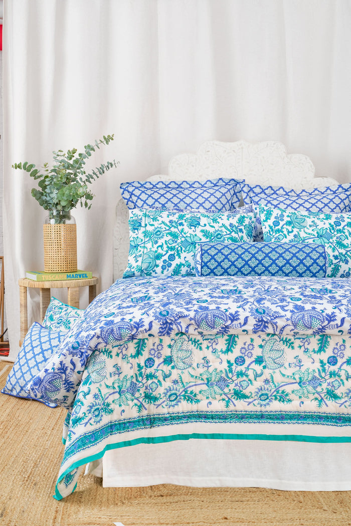 A bed styled with the Amanda Quilt by Roller Rabbit, made from 100 cotton in a blue and white floral design, sits beside a wooden nightstand holding a vase with greenery, books, and a photo frame on a woven rug against a white wall.