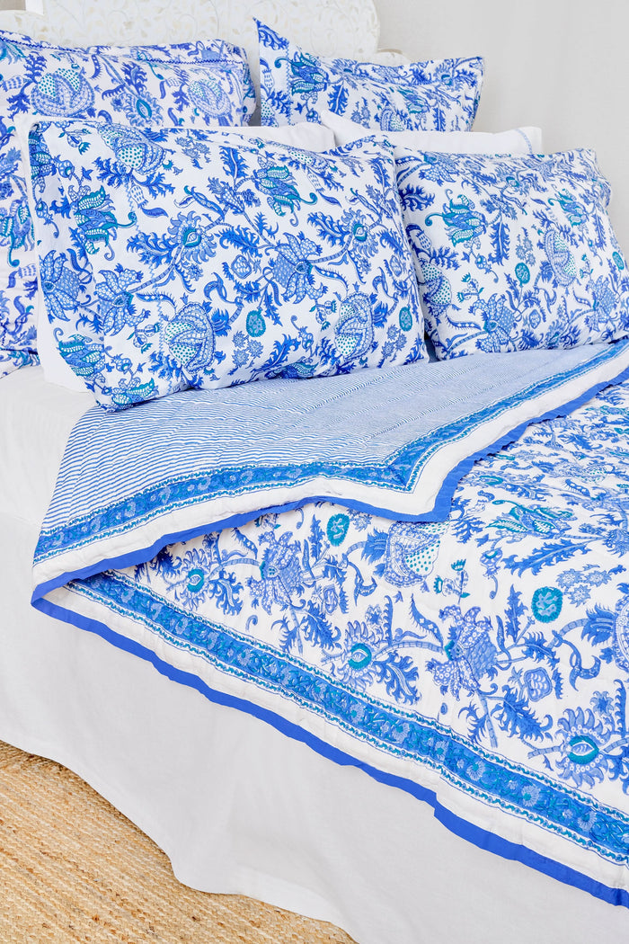 A neatly made bed displays the Roller Rabbit Amanda Quilt, crafted from 100 cotton with a blue and white floral design, set against white curtains. Beside it, a small wooden table holds books, a basket, and a vase on a woven rug.