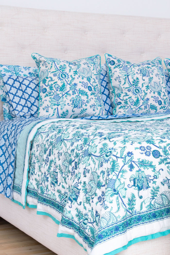 A bed styled with the Amanda Quilt by Roller Rabbit, made from 100 cotton in a blue and white floral design, sits beside a wooden nightstand holding a vase with greenery, books, and a photo frame on a woven rug against a white wall.