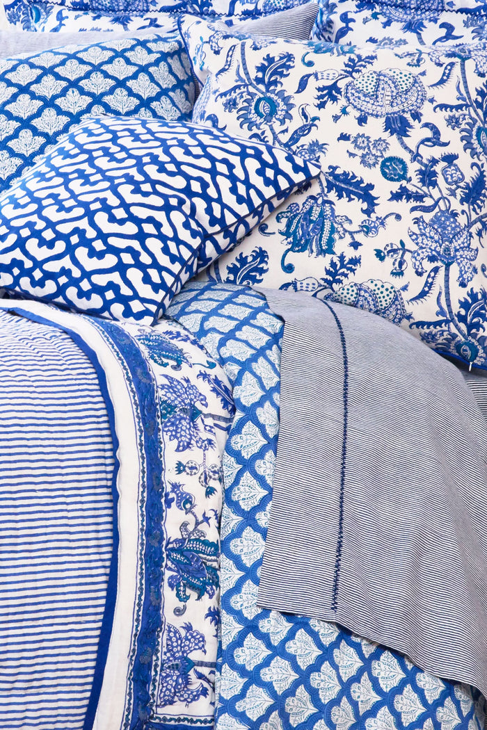 A neatly made bed displays the Roller Rabbit Amanda Quilt, crafted from 100 cotton with a blue and white floral design, set against white curtains. Beside it, a small wooden table holds books, a basket, and a vase on a woven rug.