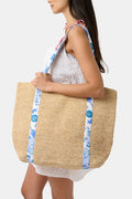 A woman in a white eyelet dress carries the Amanda Woven Beach Bag by Roller Rabbit, featuring blue and white patterned straps, over her shoulder. The cropped image shows her from shoulder to mid-thigh against a plain background.