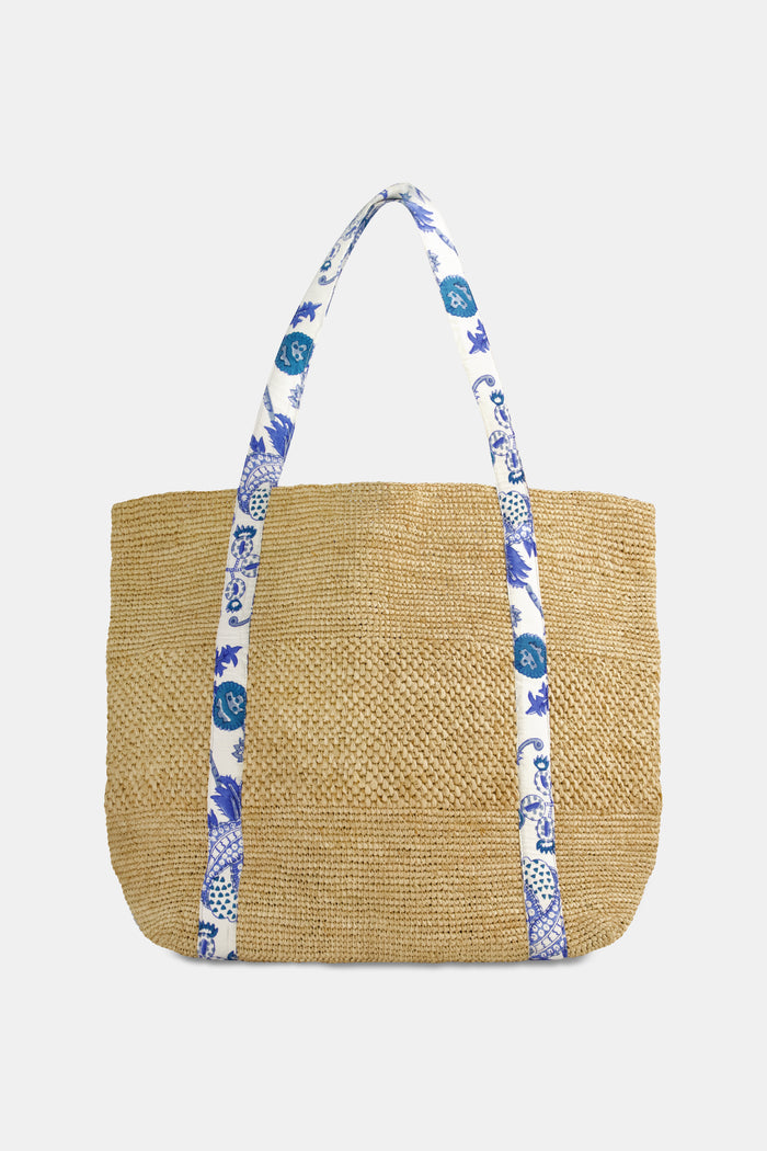 A woman in a white eyelet dress carries the Amanda Woven Beach Bag by Roller Rabbit, featuring blue and white patterned straps, over her shoulder. The cropped image shows her from shoulder to mid-thigh against a plain background.