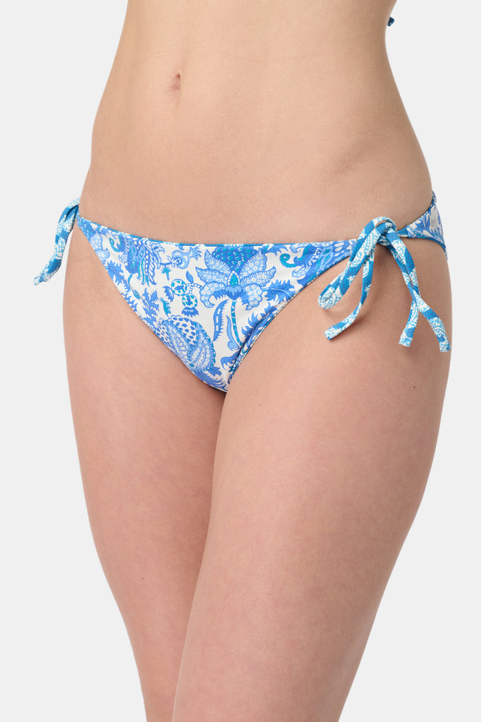 A person wears the Roller Rabbit Amanda/Jemina Serena Bikini Bottom, a reversible bikini with blue floral patterns, side ties, and UPF 50+ sun protection, standing against a plain white background.