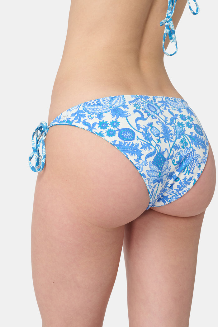 A person wears the Roller Rabbit Amanda/Jemina Serena Bikini Bottom, a reversible bikini with blue floral patterns, side ties, and UPF 50+ sun protection, standing against a plain white background.