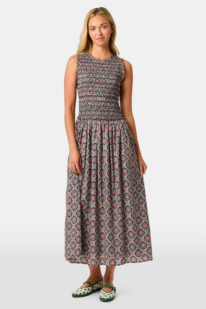 A woman wearing the Roller Rabbit Amelia Emma Dress, featuring a smocked bodice and maxi skirt, stands against a plain white background, paired with green polka-dot flats.