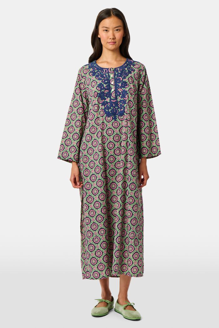 Wearing the Amelia Inez Long Kurta by Roller Rabbit, a woman with brown boots sits on the railing of a wooden porch, smiling, with a rustic building in the background.