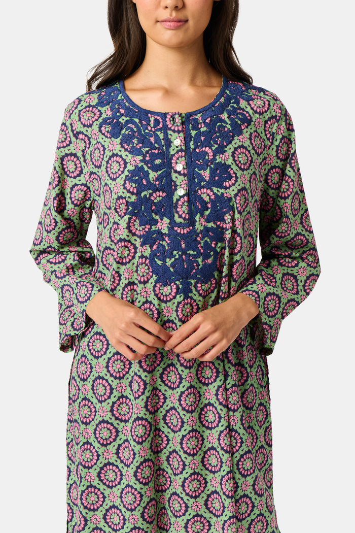 Wearing the Amelia Inez Long Kurta by Roller Rabbit, a woman with brown boots sits on the railing of a wooden porch, smiling, with a rustic building in the background.