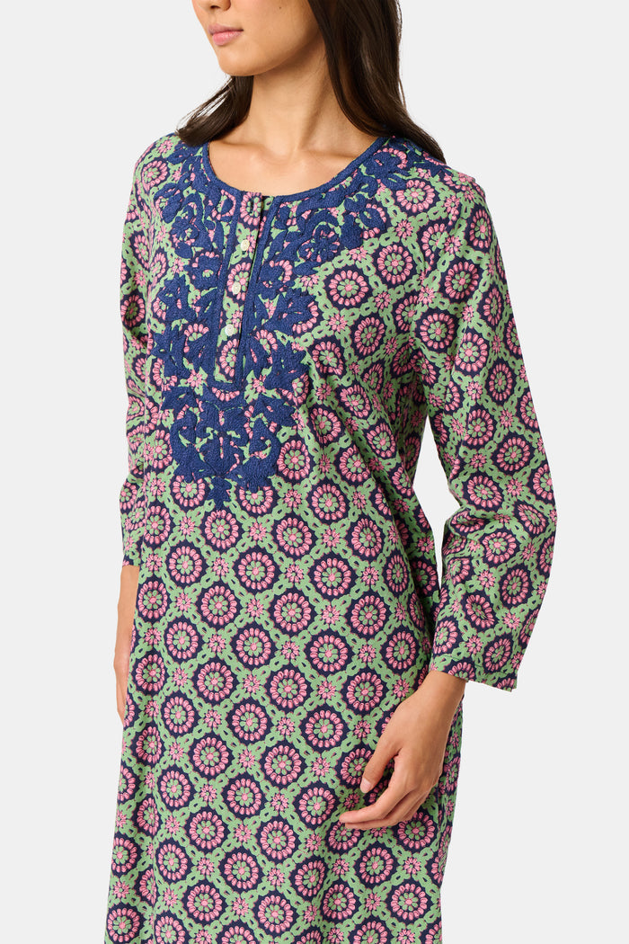 Wearing the Amelia Inez Long Kurta by Roller Rabbit, a woman with brown boots sits on the railing of a wooden porch, smiling, with a rustic building in the background.