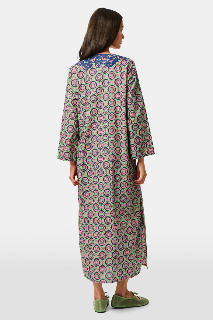 Wearing the Amelia Inez Long Kurta by Roller Rabbit, a woman with brown boots sits on the railing of a wooden porch, smiling, with a rustic building in the background.