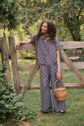 A woman stands by a wooden gate, holding a wicker basket in one hand. Outdoors amid greenery and trees, she gazes to the side with a smile, dressed in elegant Roller Rabbit Amelia Paige Pants.