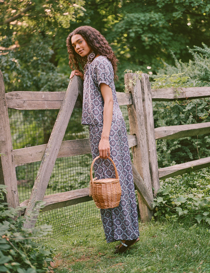 A woman stands by a wooden gate, holding a wicker basket in one hand. Outdoors amid greenery and trees, she gazes to the side with a smile, dressed in elegant Roller Rabbit Amelia Paige Pants.