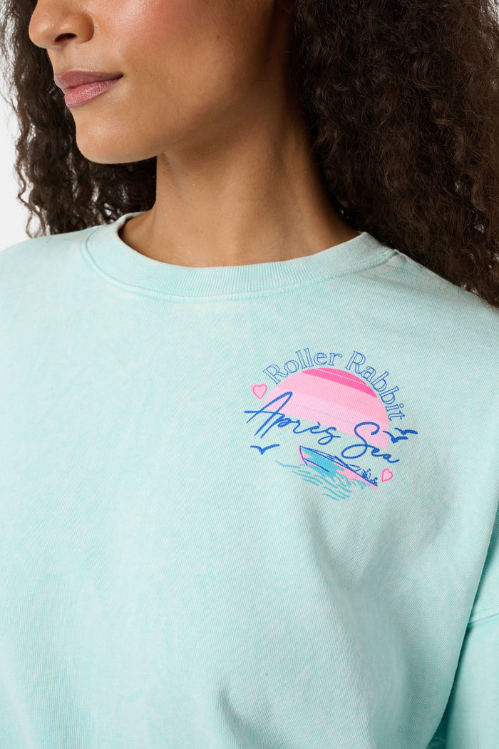 A woman wears the Roller Rabbit Apres Sea Phoebe Sweatshirt, made in the USA, mint green with a pink sunset, blue waves, birds, and "Roller Rabbit Après Sea" text on the upper left chest.