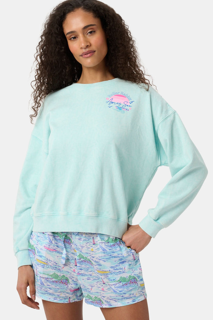 A woman wears the Roller Rabbit Apres Sea Phoebe Sweatshirt, made in the USA, mint green with a pink sunset, blue waves, birds, and "Roller Rabbit Après Sea" text on the upper left chest.