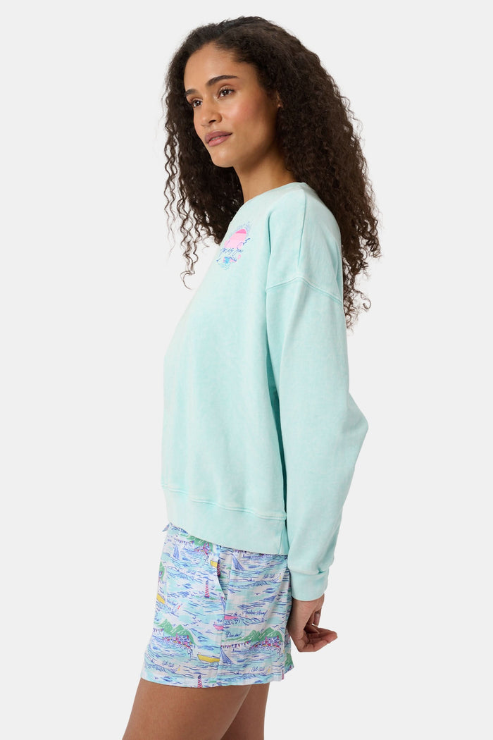 A woman wears the Roller Rabbit Apres Sea Phoebe Sweatshirt, made in the USA, mint green with a pink sunset, blue waves, birds, and "Roller Rabbit Après Sea" text on the upper left chest.