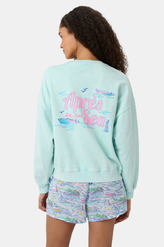 A woman wears the Roller Rabbit Apres Sea Phoebe Sweatshirt, made in the USA, mint green with a pink sunset, blue waves, birds, and "Roller Rabbit Après Sea" text on the upper left chest.