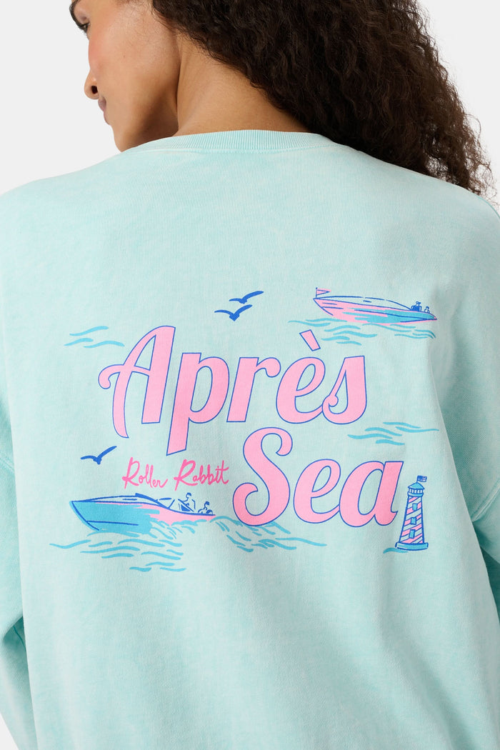 A woman wears the Roller Rabbit Apres Sea Phoebe Sweatshirt, made in the USA, mint green with a pink sunset, blue waves, birds, and "Roller Rabbit Après Sea" text on the upper left chest.