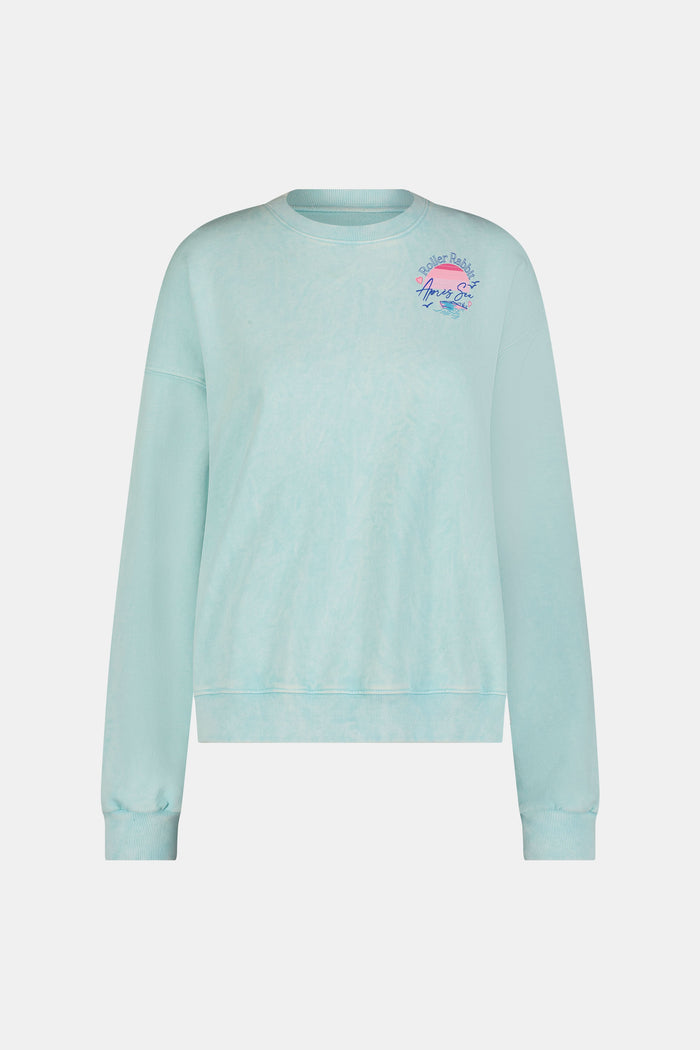 A woman wears the Roller Rabbit Apres Sea Phoebe Sweatshirt, made in the USA, mint green with a pink sunset, blue waves, birds, and "Roller Rabbit Après Sea" text on the upper left chest.