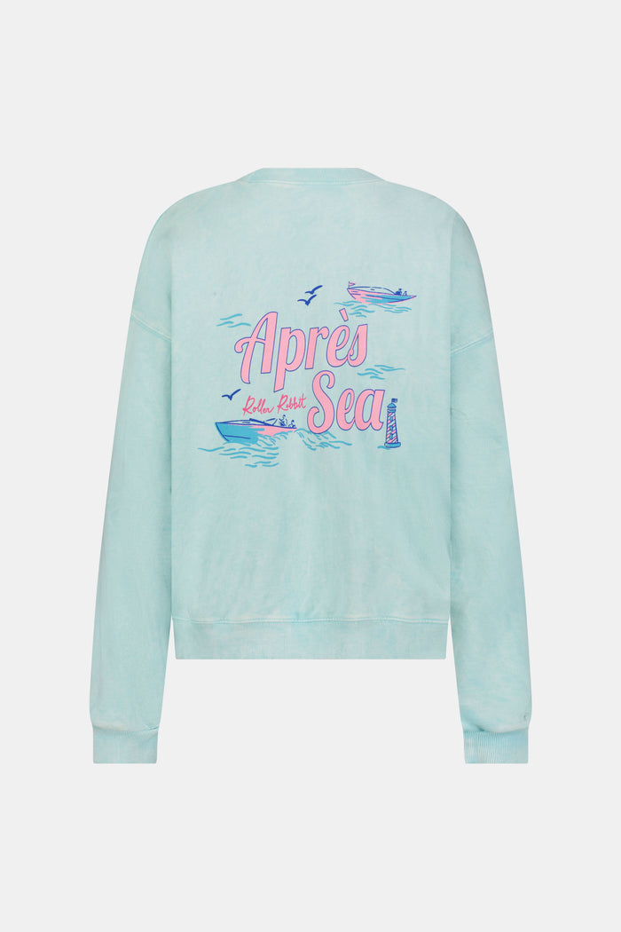 A woman wears the Roller Rabbit Apres Sea Phoebe Sweatshirt, made in the USA, mint green with a pink sunset, blue waves, birds, and "Roller Rabbit Après Sea" text on the upper left chest.