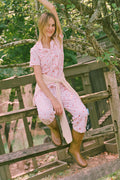A woman in the Roller Rabbit Autumn Hearts Fiora Pajamas and brown boots sits on a wooden fence, smiling and resting one arm on a tree branch in a sunny outdoor setting, evoking the coziness of fall sleepwear.