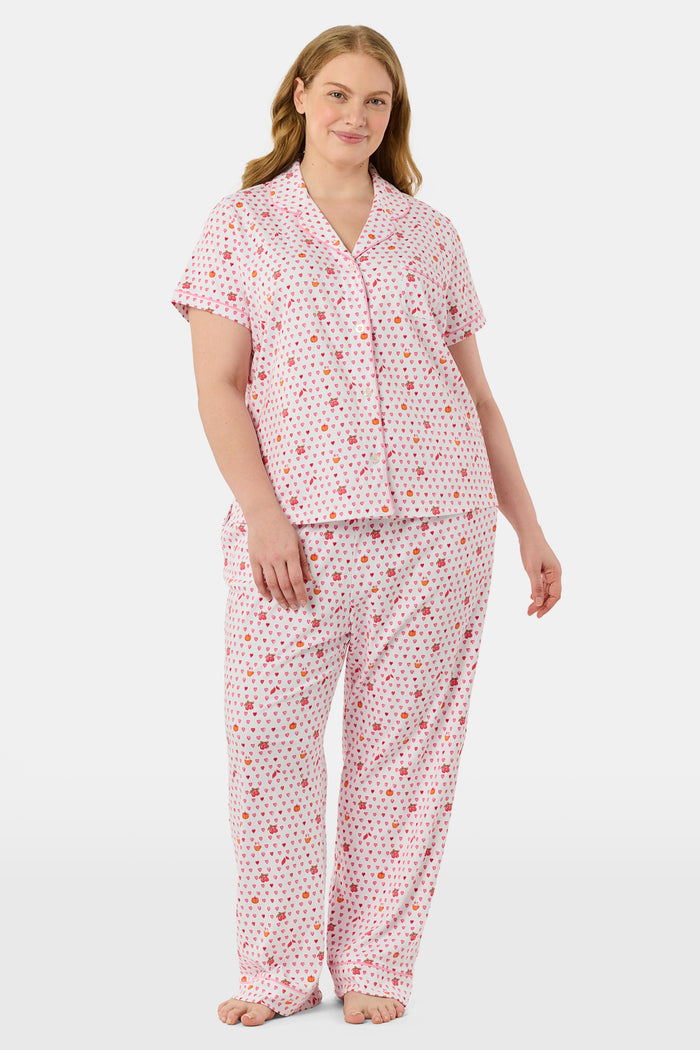 A woman in the Roller Rabbit Autumn Hearts Fiora Pajamas and brown boots sits on a wooden fence, smiling and resting one arm on a tree branch in a sunny outdoor setting, evoking the coziness of fall sleepwear.
