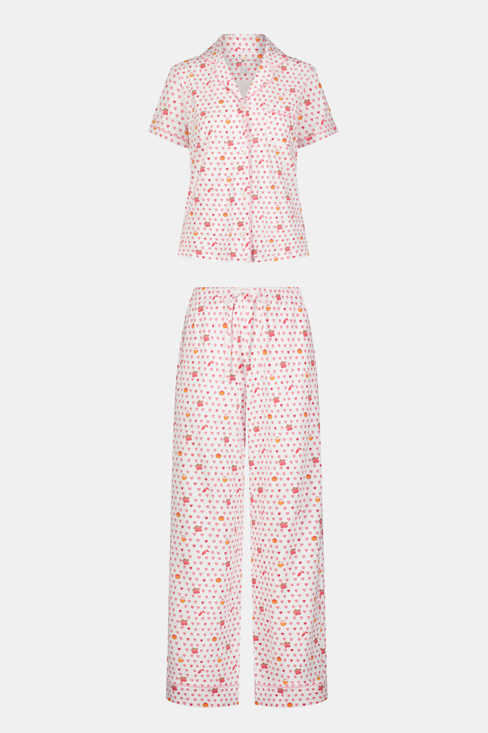 A woman in the Roller Rabbit Autumn Hearts Fiora Pajamas and brown boots sits on a wooden fence, smiling and resting one arm on a tree branch in a sunny outdoor setting, evoking the coziness of fall sleepwear.