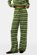 A person wearing the Roller Rabbit Autumn Stripe Olivia Pant—green and white striped, pull-on, flared—and a matching sweater lifts the hem to reveal their midriff. Brown slippers are visible. The background is white.
