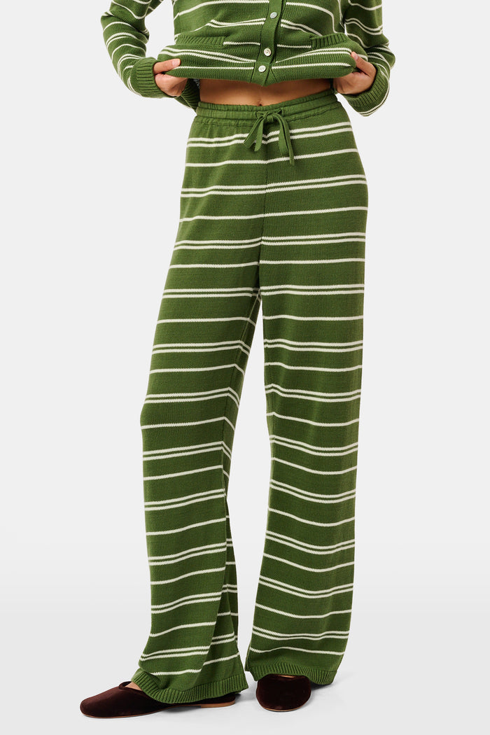 A person wearing the Roller Rabbit Autumn Stripe Olivia Pant—green and white striped, pull-on, flared—and a matching sweater lifts the hem to reveal their midriff. Brown slippers are visible. The background is white.
