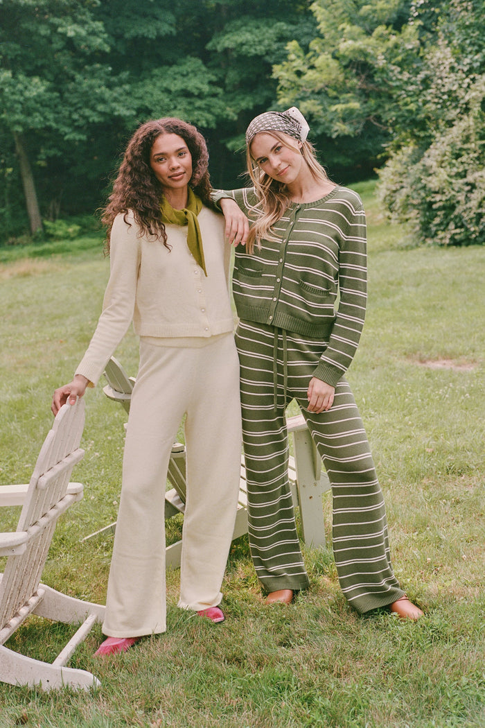A person wearing the Roller Rabbit Autumn Stripe Olivia Pant—green and white striped, pull-on, flared—and a matching sweater lifts the hem to reveal their midriff. Brown slippers are visible. The background is white.