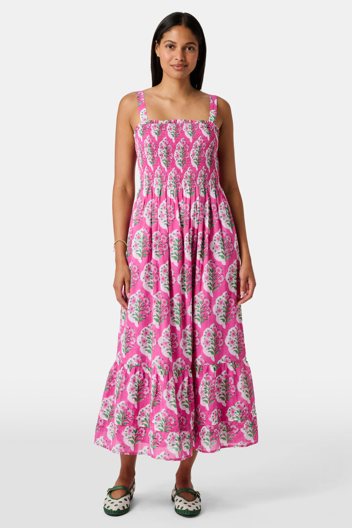 The Kids Ava Floral Ceri Dress by Roller Rabbit is a sleeveless baby dress featuring a smocked bodice and ruffled straps, adorned with green and white leaf patterns on a vibrant pink background, and comes with matching bloomers.