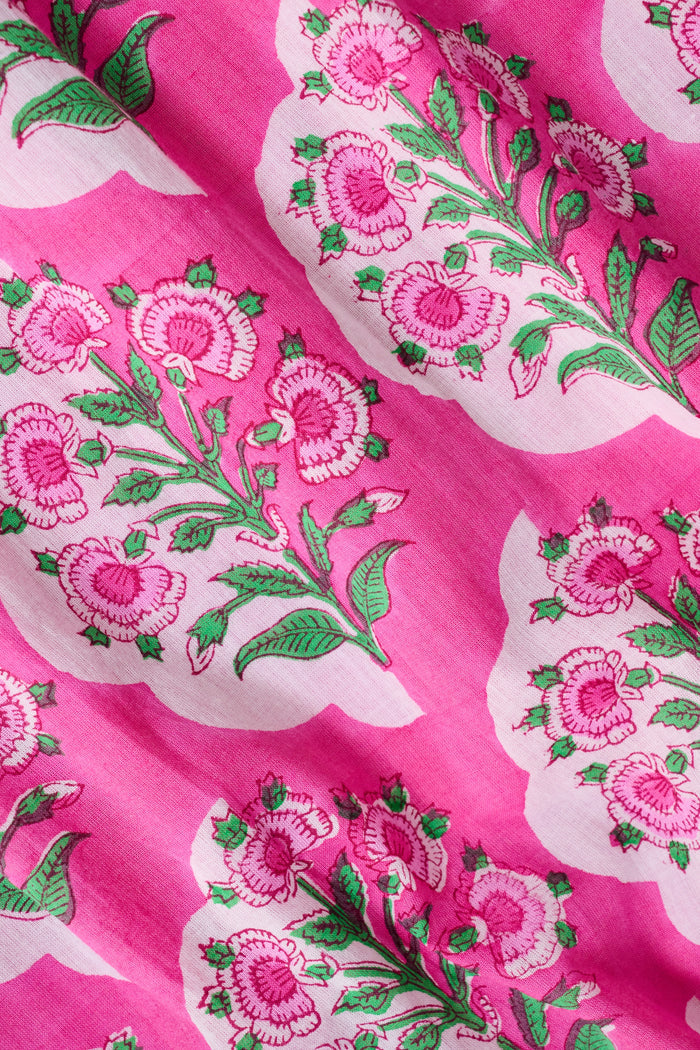 A person wearing the Roller Rabbit Ava Floral Bristol Dress—a pink cotton voile dress with a smocked bodice—has a green sweater draped over their shoulders and stands in a library near bookshelves.