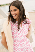A woman with long brown hair smiles outdoors, wearing the Roller Rabbit Big Cata Pippa Dress—a short-sleeved pink cotton voile dress with a geometric pattern—paired with a light sweater. Sandy beach and water form the background.