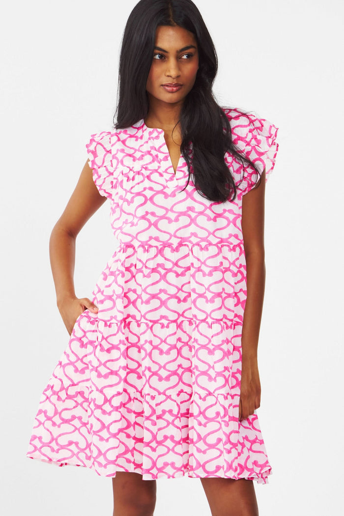 A woman with long brown hair smiles outdoors, wearing the Roller Rabbit Big Cata Pippa Dress—a short-sleeved pink cotton voile dress with a geometric pattern—paired with a light sweater. Sandy beach and water form the background.