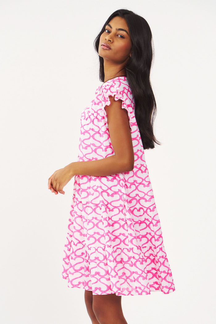 A woman with long brown hair smiles outdoors, wearing the Roller Rabbit Big Cata Pippa Dress—a short-sleeved pink cotton voile dress with a geometric pattern—paired with a light sweater. Sandy beach and water form the background.