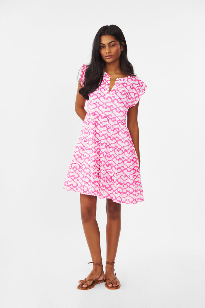 A woman with long brown hair smiles outdoors, wearing the Roller Rabbit Big Cata Pippa Dress—a short-sleeved pink cotton voile dress with a geometric pattern—paired with a light sweater. Sandy beach and water form the background.