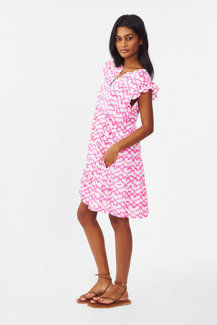 A woman with long brown hair smiles outdoors, wearing the Roller Rabbit Big Cata Pippa Dress—a short-sleeved pink cotton voile dress with a geometric pattern—paired with a light sweater. Sandy beach and water form the background.