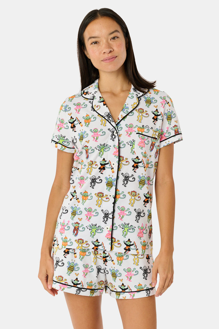 A smiling child in Roller Rabbit’s Kids Boo Crew Pajamas, crafted from soft Pima cotton, sits with arms raised on outdoor steps among women in coordinating patterned pajamas, all sharing a cheerful and relaxed moment together.