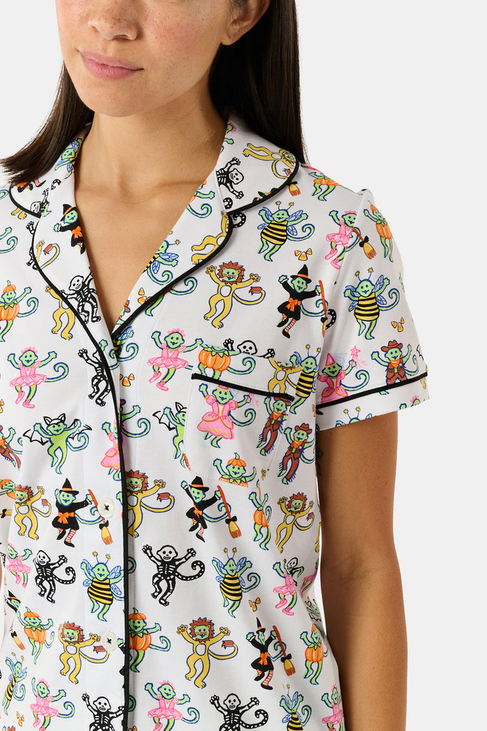 A smiling child in Roller Rabbit’s Kids Boo Crew Pajamas, crafted from soft Pima cotton, sits with arms raised on outdoor steps among women in coordinating patterned pajamas, all sharing a cheerful and relaxed moment together.