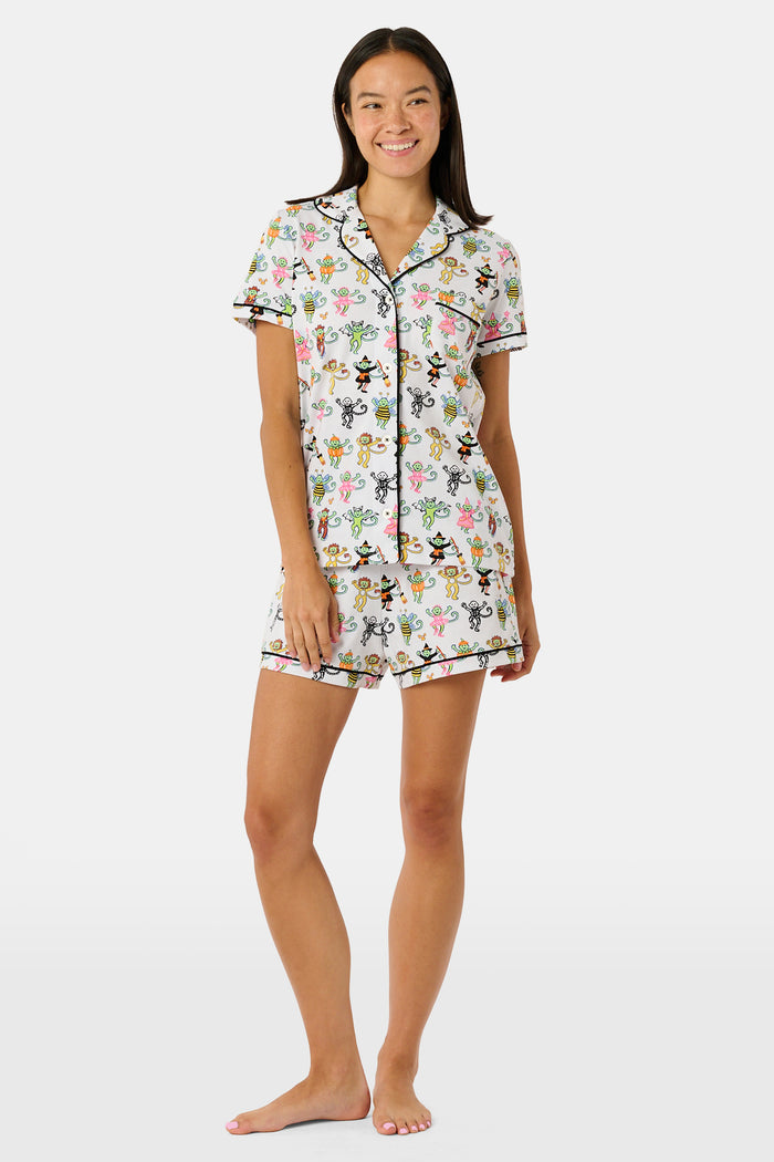 A smiling child in Roller Rabbit’s Kids Boo Crew Pajamas, crafted from soft Pima cotton, sits with arms raised on outdoor steps among women in coordinating patterned pajamas, all sharing a cheerful and relaxed moment together.