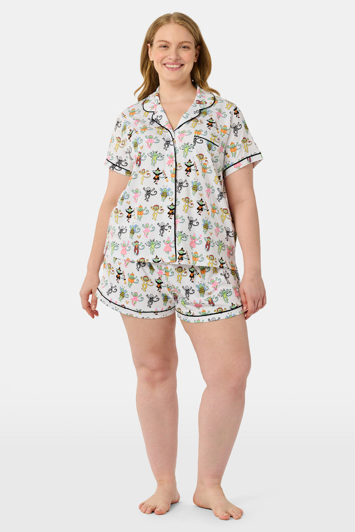 A smiling child in Roller Rabbit’s Kids Boo Crew Pajamas, crafted from soft Pima cotton, sits with arms raised on outdoor steps among women in coordinating patterned pajamas, all sharing a cheerful and relaxed moment together.