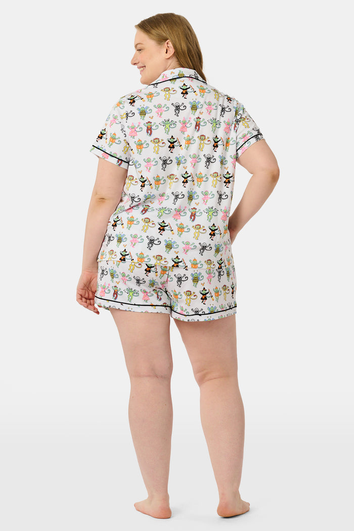 A smiling child in Roller Rabbit’s Kids Boo Crew Pajamas, crafted from soft Pima cotton, sits with arms raised on outdoor steps among women in coordinating patterned pajamas, all sharing a cheerful and relaxed moment together.
